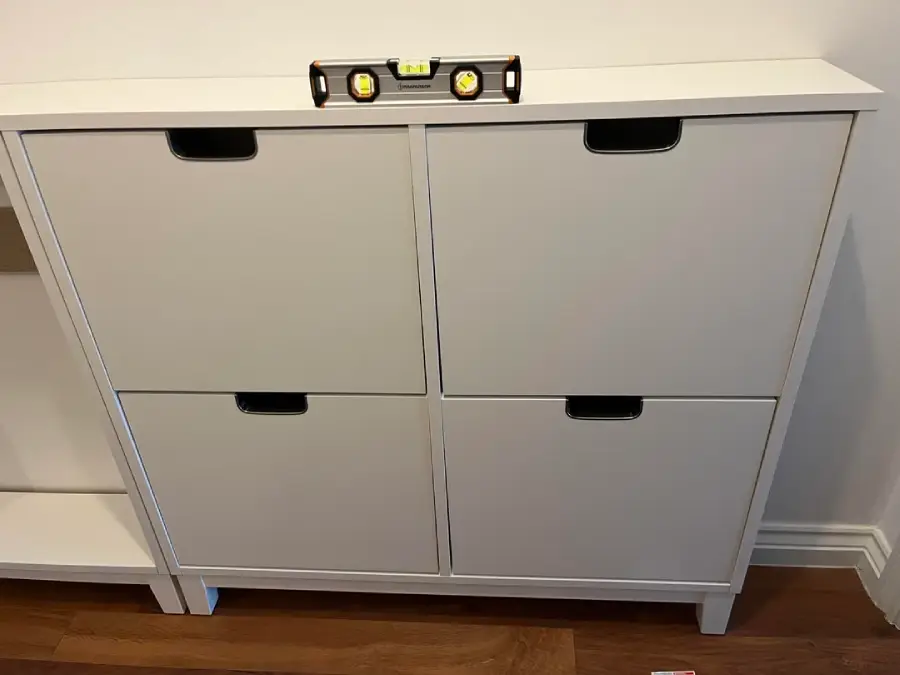 A white cabinet with four compartments stands on a wooden floor. A spirit level is placed on top to check alignment. The wall behind is plain and white.