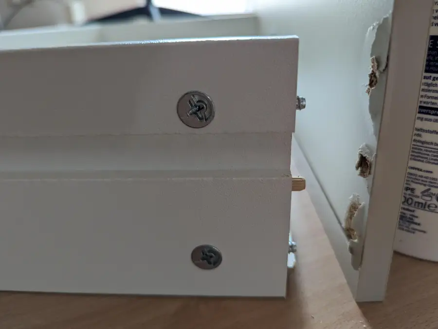 Close-up of white furniture assembly showing mismatched screws and dowel; the side panel’s holes do not align with the connected pieces, causing improper fit.