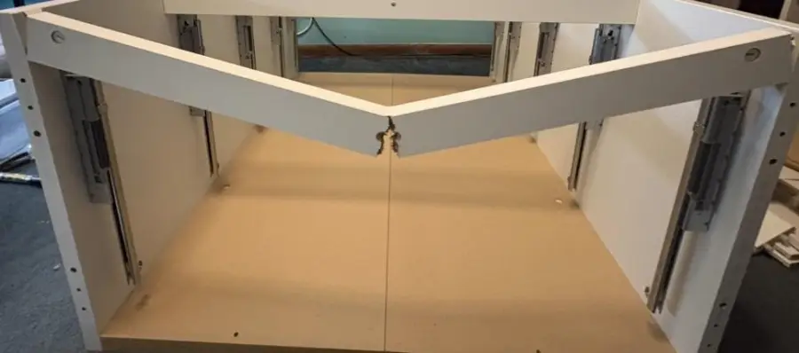 A partially assembled cabinet with a broken central support beam, showing a large crack in the middle. The unit is white with metal drawer slides on the sides, placed on a flat surface indoors.