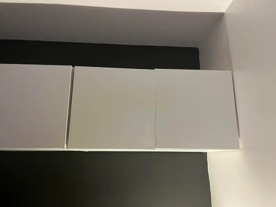 Three white rectangular cabinet doors are mounted on a dark wall, viewed from below at an angle. The lighting casts soft shadows, accentuating the minimalist design and clean lines of the cabinets and surrounding surfaces.