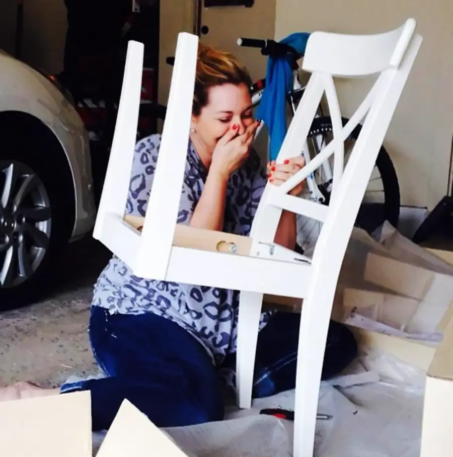 A woman sits on the floor laughing while holding a white chair upside down, possibly assembling it, in a garage with a car and bicycle in the background.