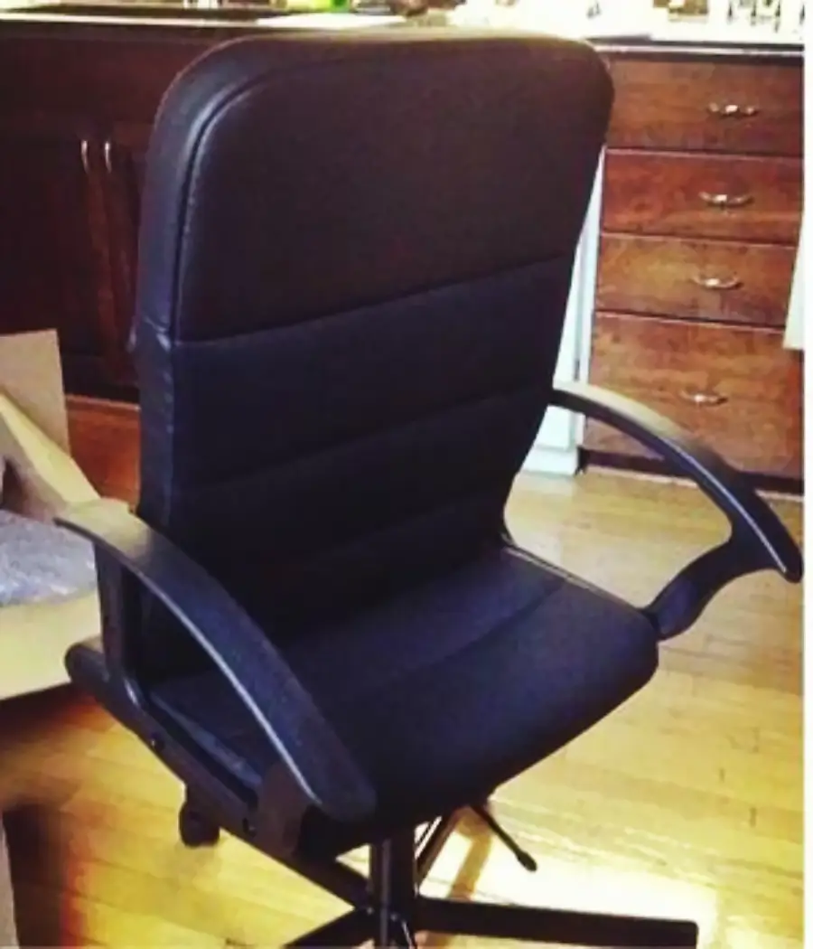 A black office chair with padded seat and backrest sits on a wooden floor near a set of wooden drawers and kitchen cabinets.