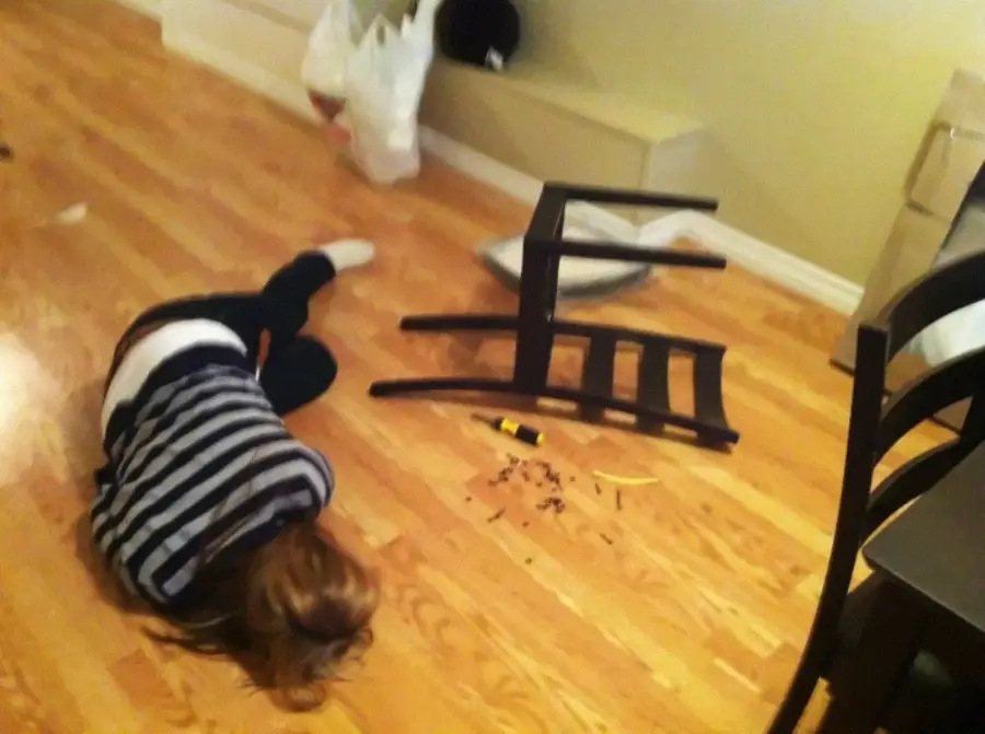 A person lies on a wooden floor next to an overturned chair, a screwdriver, and scattered screws, suggesting they may have fallen while assembling furniture.