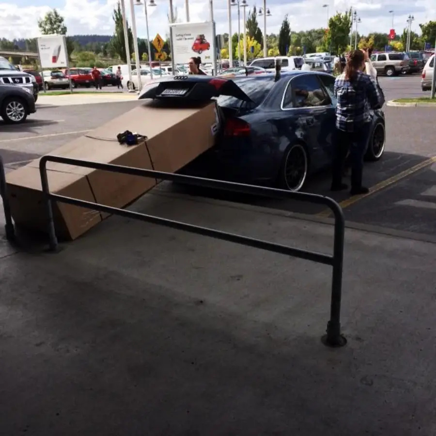 A large cardboard box sticks far out of the trunk of a parked car while two people stand nearby in a parking lot. The box extends over a nearby barrier, blocking part of the walkway.