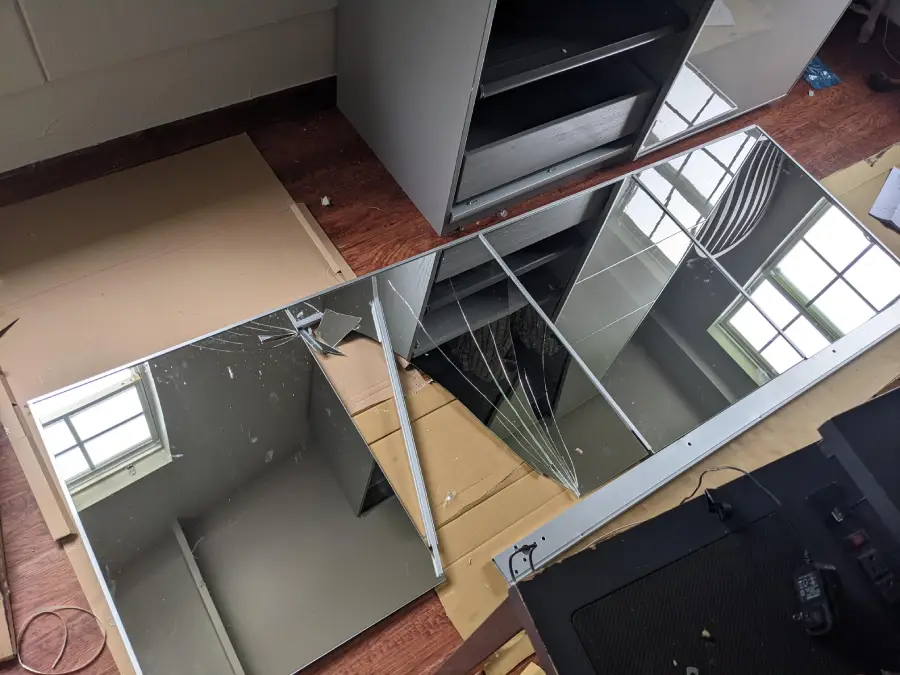 A large rectangular mirror is lying on the floor, cracked and broken in several places, reflecting windows and part of the room. There are cabinets and wooden boards nearby.