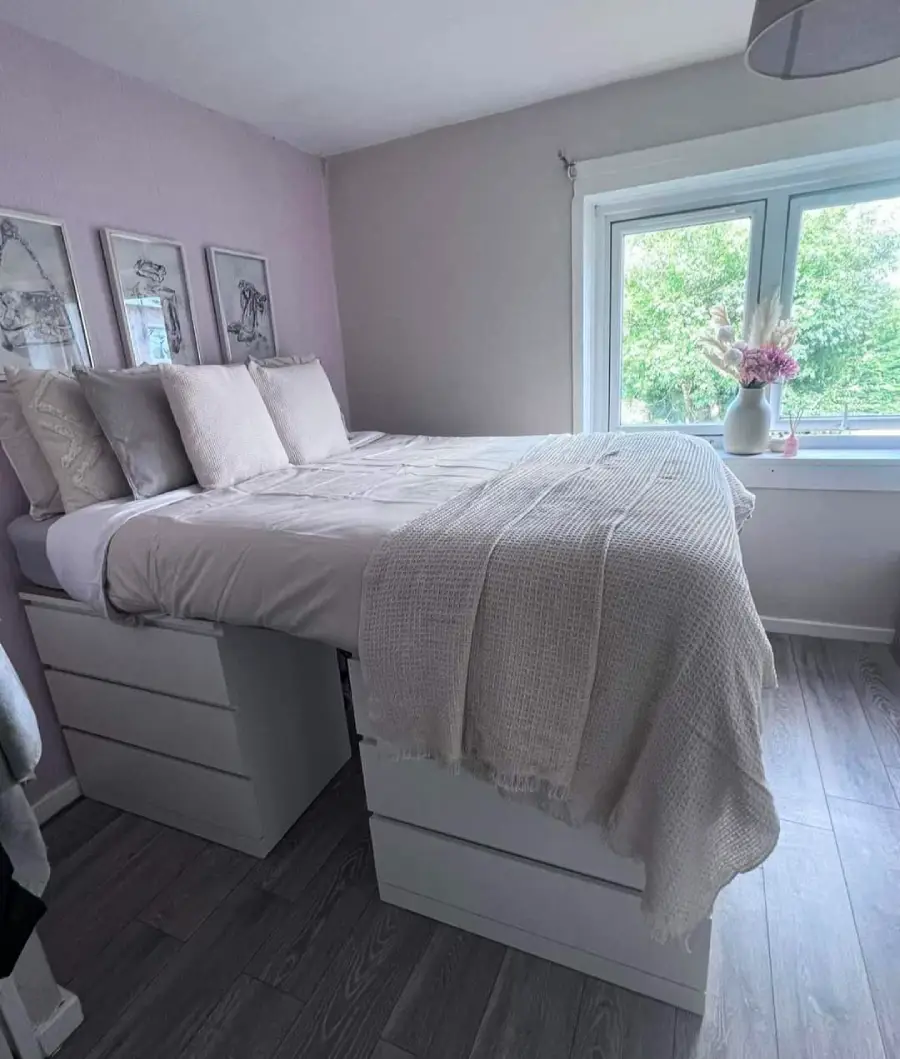 A bed with neutral bedding is elevated on four white dresser units in a cozy bedroom; a window with a vase of flowers and wall art are visible.