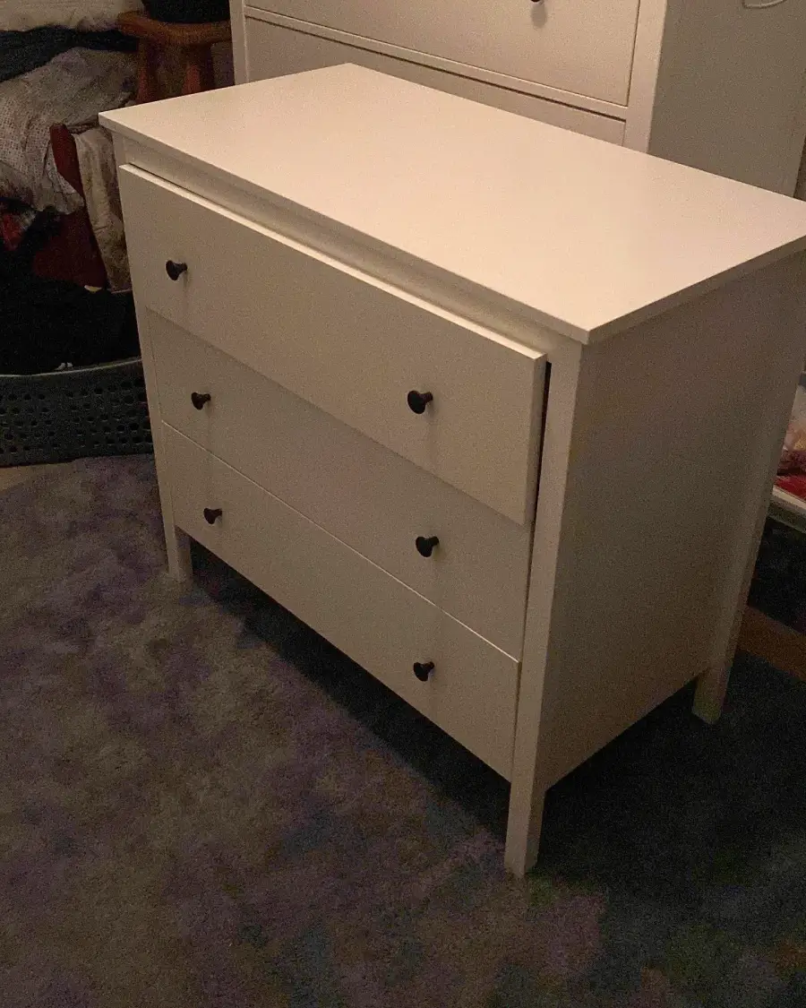A white three-drawer dresser with heart-shaped black knobs stands on a carpeted floor, with other furniture and items visible in the dimly lit background.