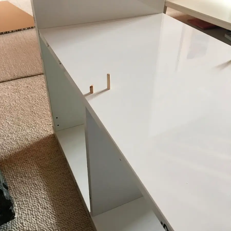 White, glossy furniture panel with two wooden dowels sticking out, partially assembled on a beige carpet, surrounded by other boards and parts.