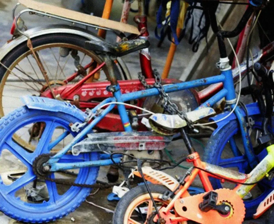 Three dusty, chained bicycles—one blue, one red, and one orange—are clustered together in a cluttered area, showing visible wear and tear.