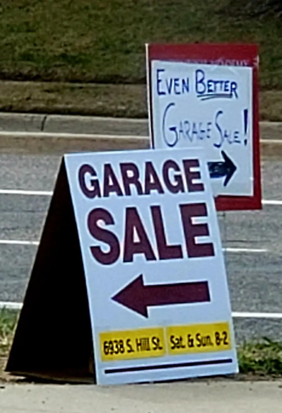 A sandwich board sign reads “GARAGE SALE” with a left arrow and details below. Behind it, another sign says “Even Better Garage Sale!” with a right arrow. Both are by a roadside.