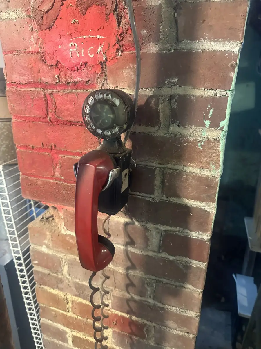 A vintage rotary payphone with a red handset is mounted on a worn brick wall. The phone is old-fashioned with a coiled cord hanging down.