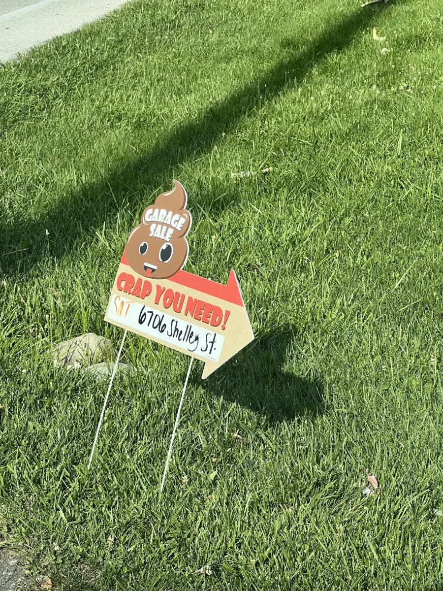 A garage sale sign shaped like a poop emoji with “Grab you need!” and an arrow pointing right, displaying the address “6706 Shelley St.,” is staked into green grass beside a sidewalk.