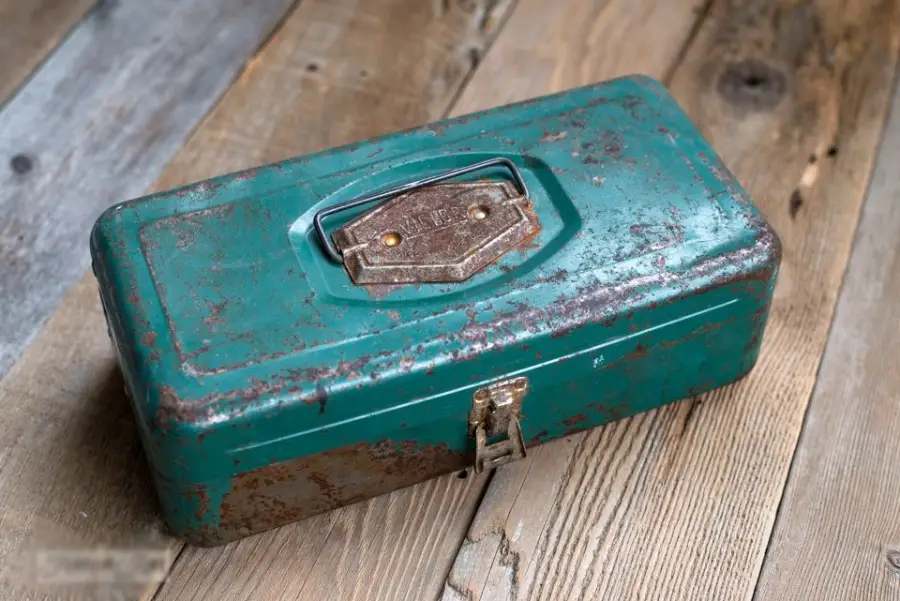 A weathered, green metal cash box with rust spots sits closed on a wooden surface, showing a handle and latch on top.