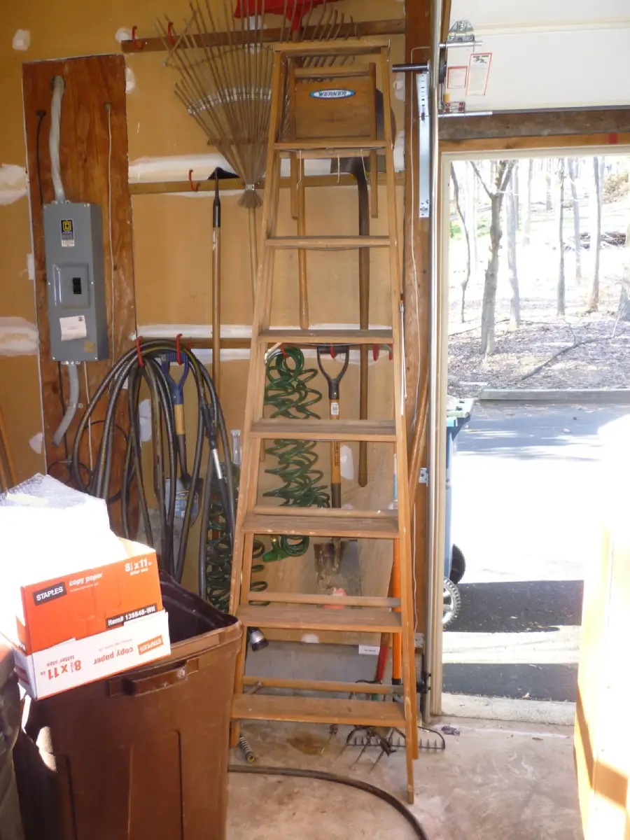 A wooden ladder stands in a garage with rakes, hoses, boxes, and garden tools on the walls and floor. Sunlight shines through an open door, revealing trees outside.