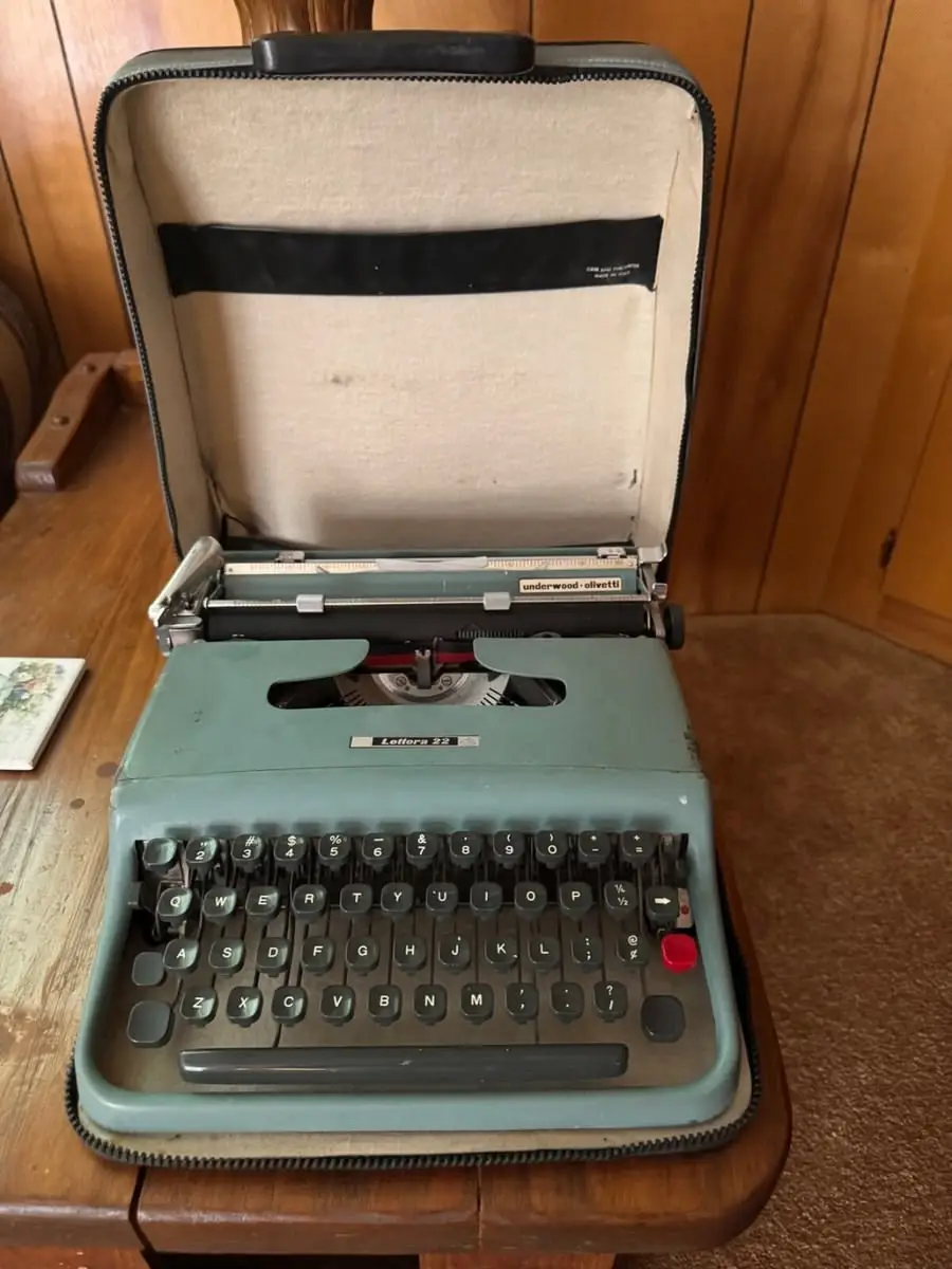 A vintage blue portable typewriter with black keys sits open in its carrying case on a wooden table, against a wood-paneled wall.