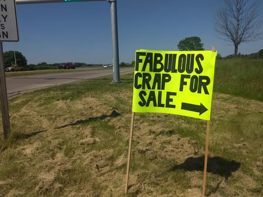 A bright yellow roadside sign with bold black text reads “FABULOUS CRAP FOR SALE” and points to the right. The sign is on a grassy area beside a road under a clear blue sky.