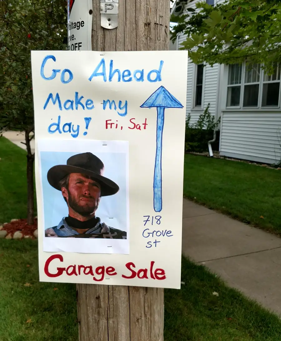Handwritten garage sale sign on a pole features a photo of Clint Eastwood, blue text saying “Go ahead make my day!” with a blue arrow, and red “Garage Sale” at the bottom. Includes address: 718 Grove St, Fri, Sat.