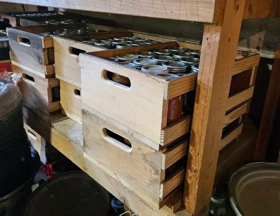 Wooden crates stacked on shelves, each crate filled with glass jars, likely used for home canning or storage. The setting appears to be a pantry or storage room.