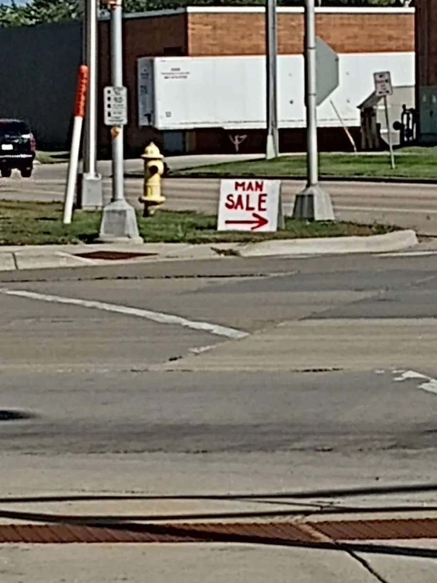 A handwritten sign on grass by a street reads “MAN SALE” with a red arrow pointing right.