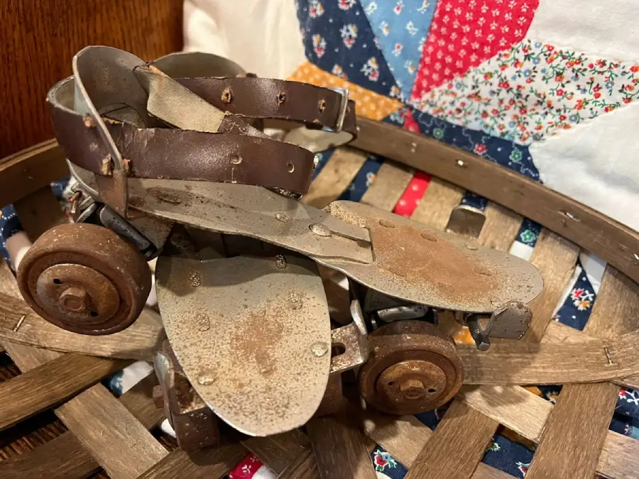 A pair of vintage metal roller skates with brown leather straps sit on a woven basket, with a colorful patchwork quilt in the background.