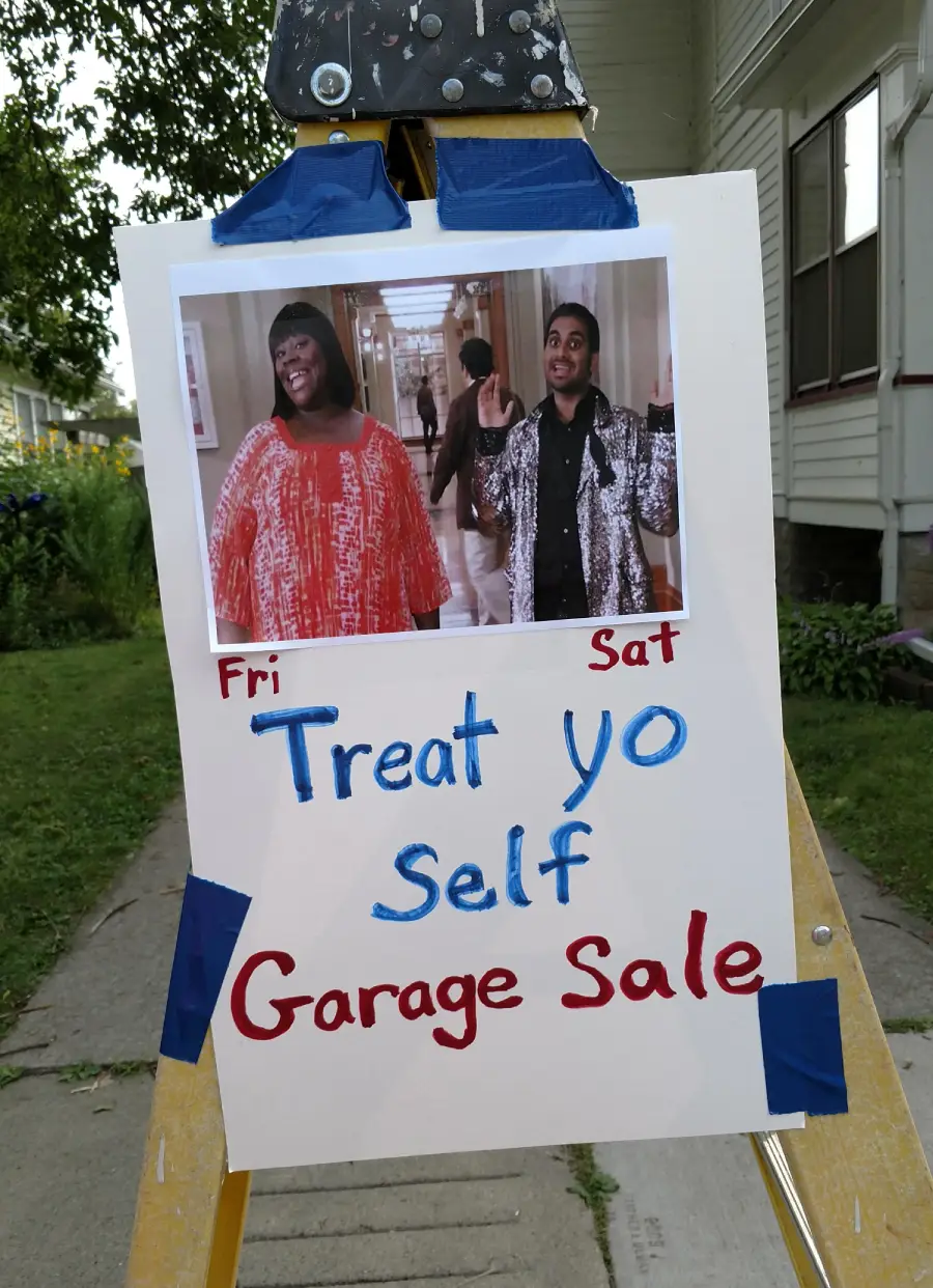 A sidewalk sign with photos of two smiling people, text reads: “Fri Sat Treat Yo Self Garage Sale,” promoting a weekend garage sale. The sign is taped to an easel outside a house.