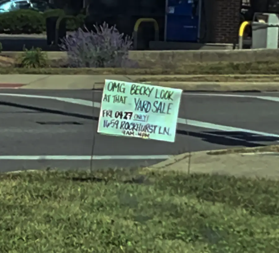 A handwritten yard sale sign on a lawn reads, “OMG BECKY LOOK AT THAT YARD SALE. Fri 09/22 only, 1159 Brookhurst Ln.” It’s posted near a sidewalk and street.
