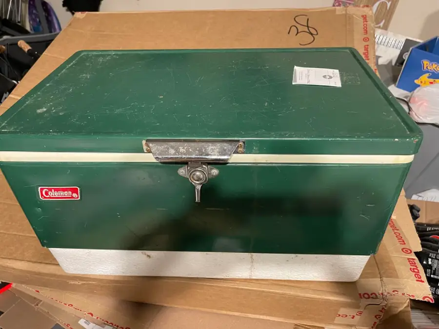 A green and white vintage Coleman cooler sits on a cardboard box, with a metal latch and a small white sticker on top.