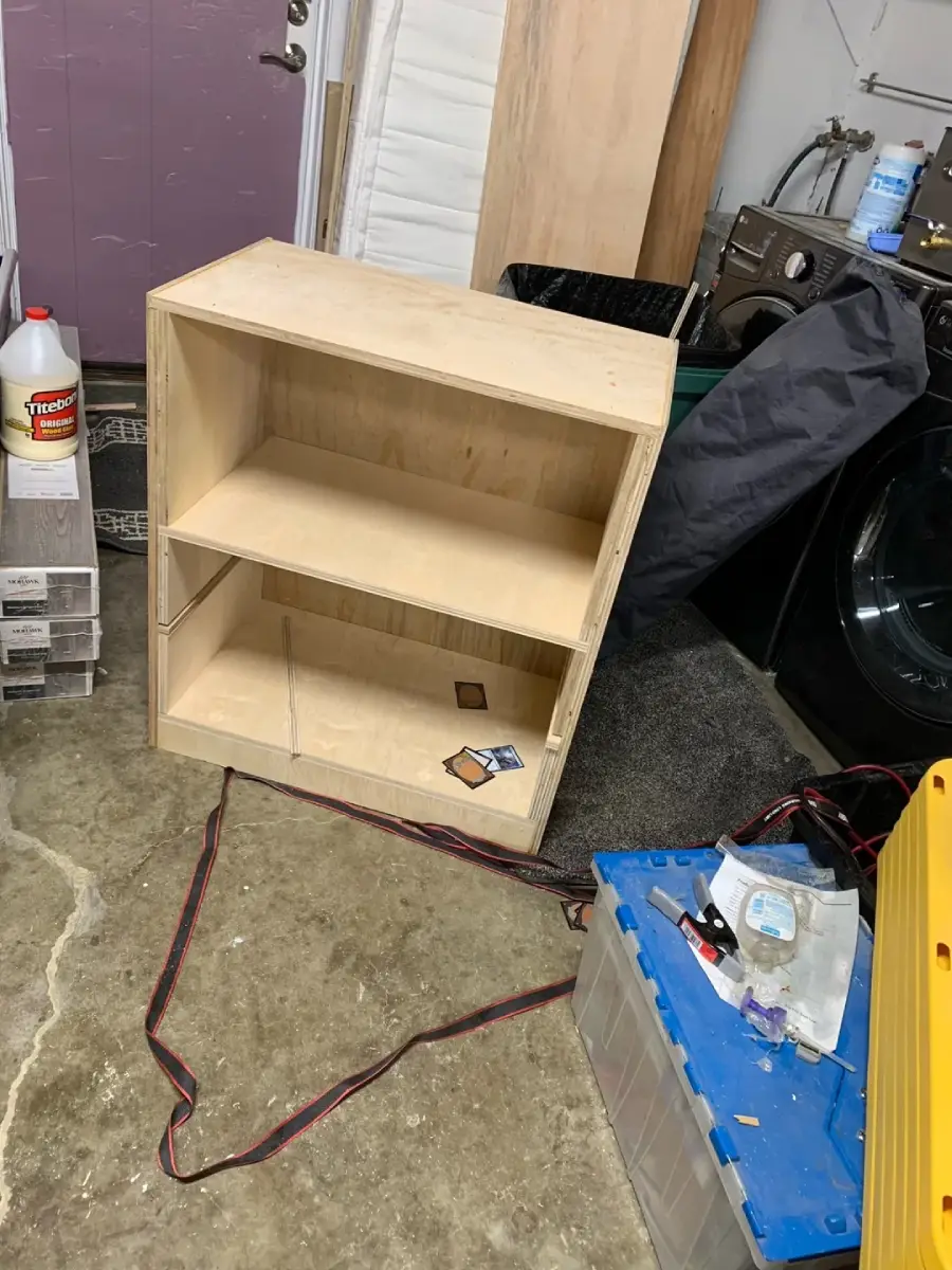 A wooden shelf stands on a concrete floor in a cluttered room with tools, bottles, and a washing machine nearby. Two SD cards are on one shelf.