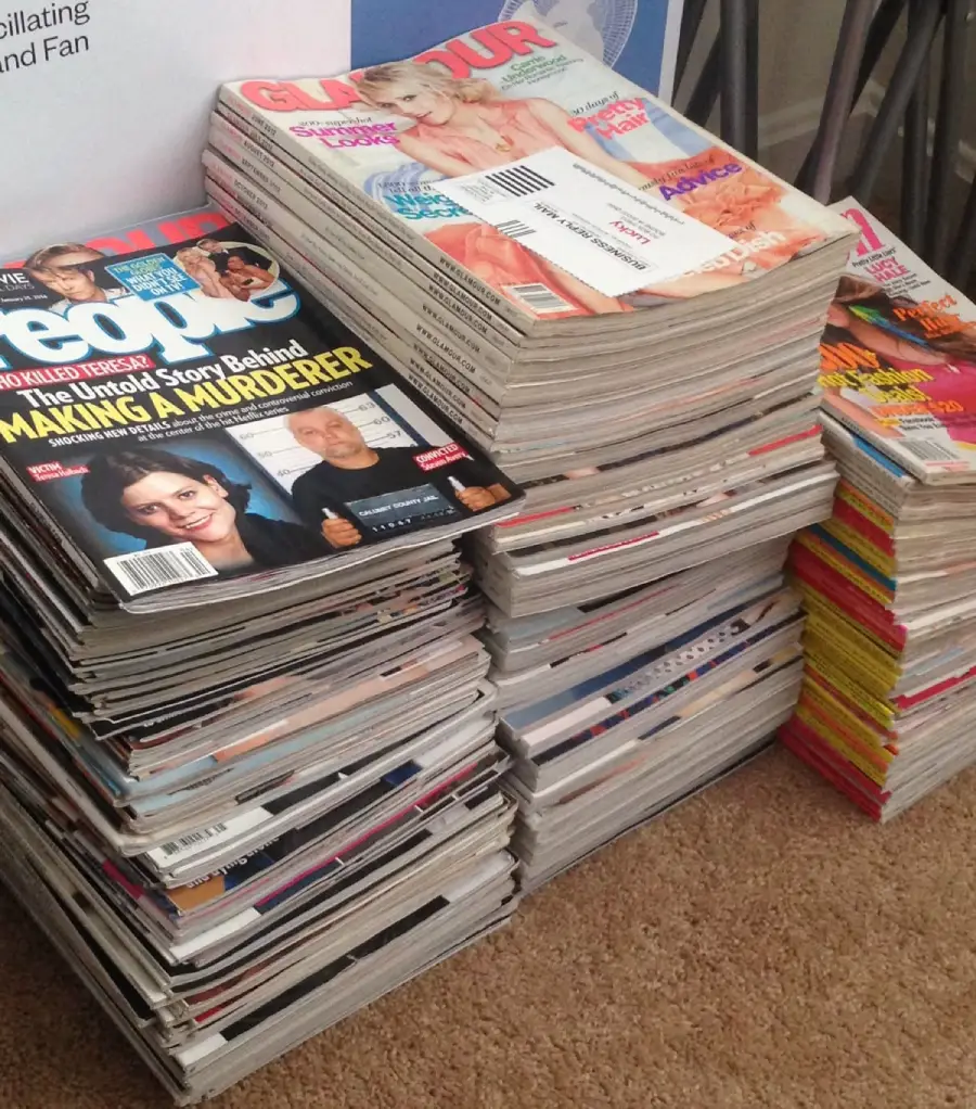 Three tall stacks of assorted magazines, including “People” and “Glamour,” are arranged on a carpeted floor. Some magazines feature celebrities on their covers, and one has a visible mailing label.