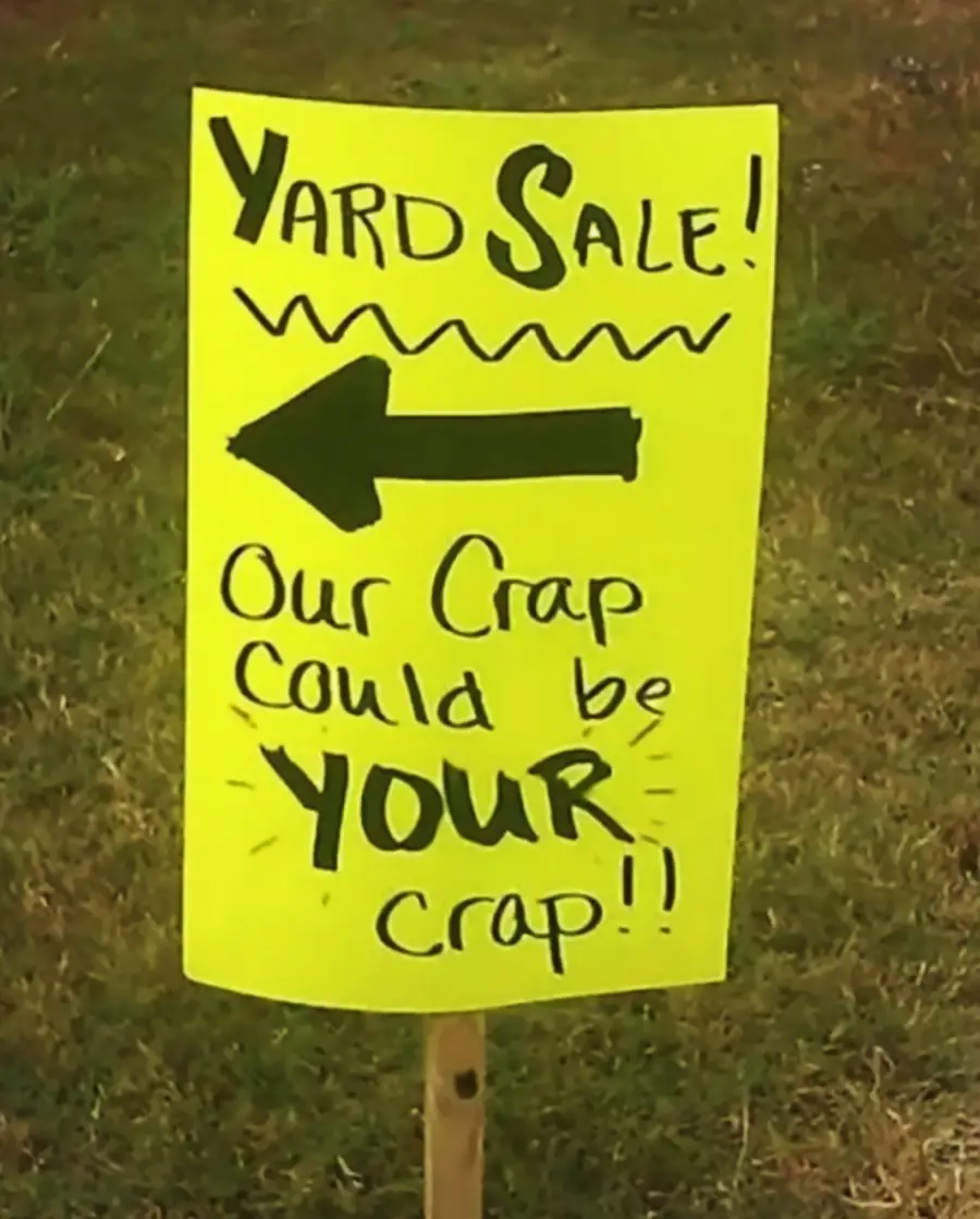 A bright yellow yard sale sign with a left-pointing arrow reads: “Yard Sale! Our crap could be YOUR crap!!” The sign is placed on grass.