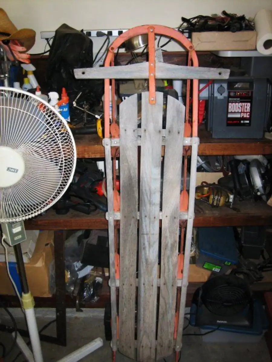 A vintage wooden sled with red metal runners leans against a cluttered workshop shelf; a white floor fan is on the left and various tools and boxes are visible in the background.
