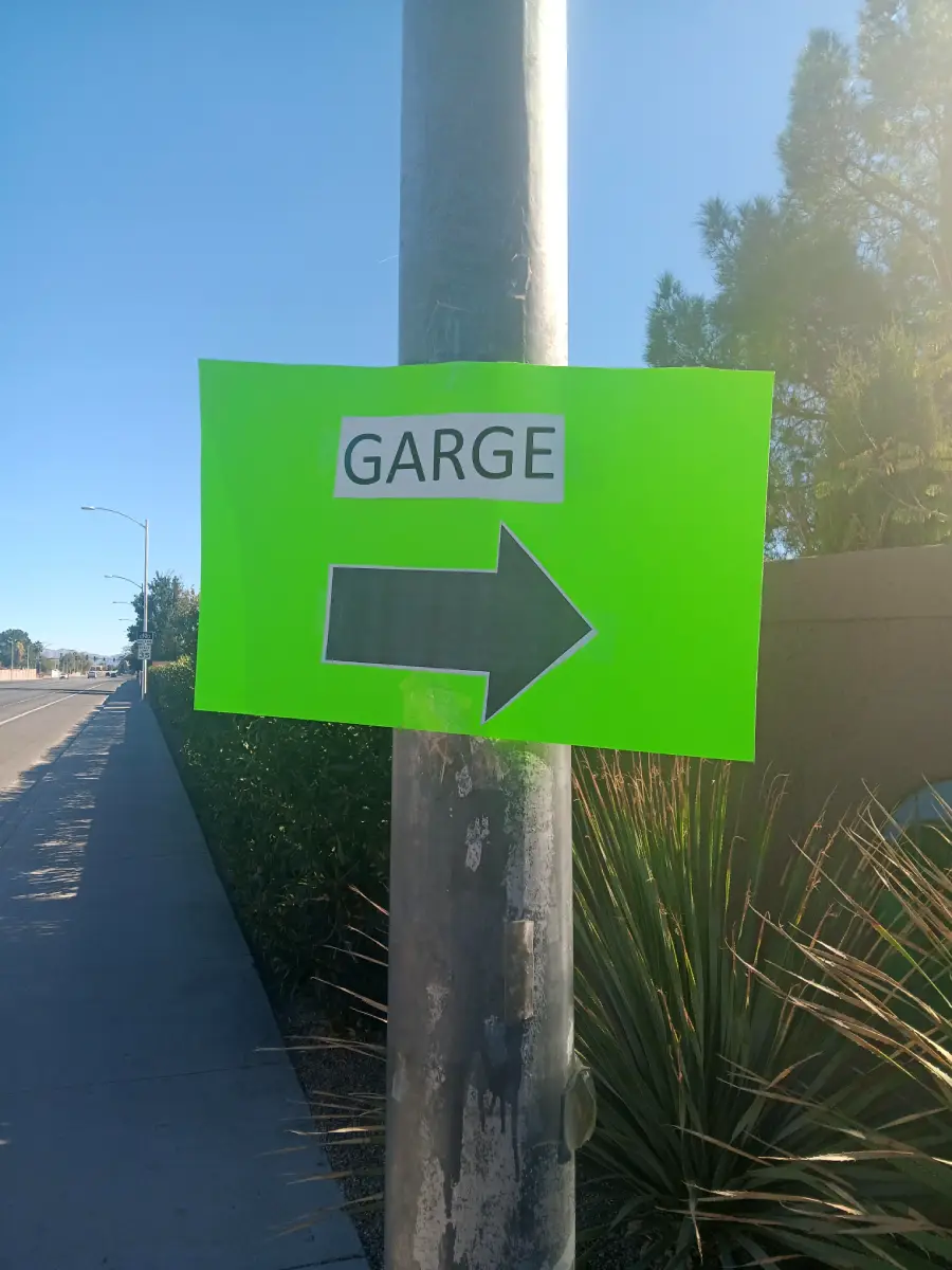 A bright green sign with the word “GARGE” and a large black arrow pointing right is taped to a pole along a sidewalk.