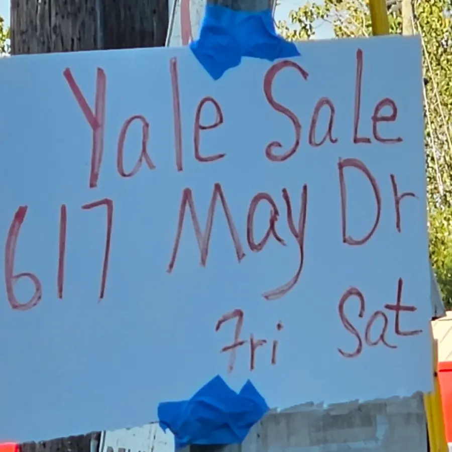 Handwritten sign on white paper with red marker reads: “Yale Sale, 617 May Dr, Fri Sat.” The sign is taped to a surface with blue tape, and trees are visible in the background.