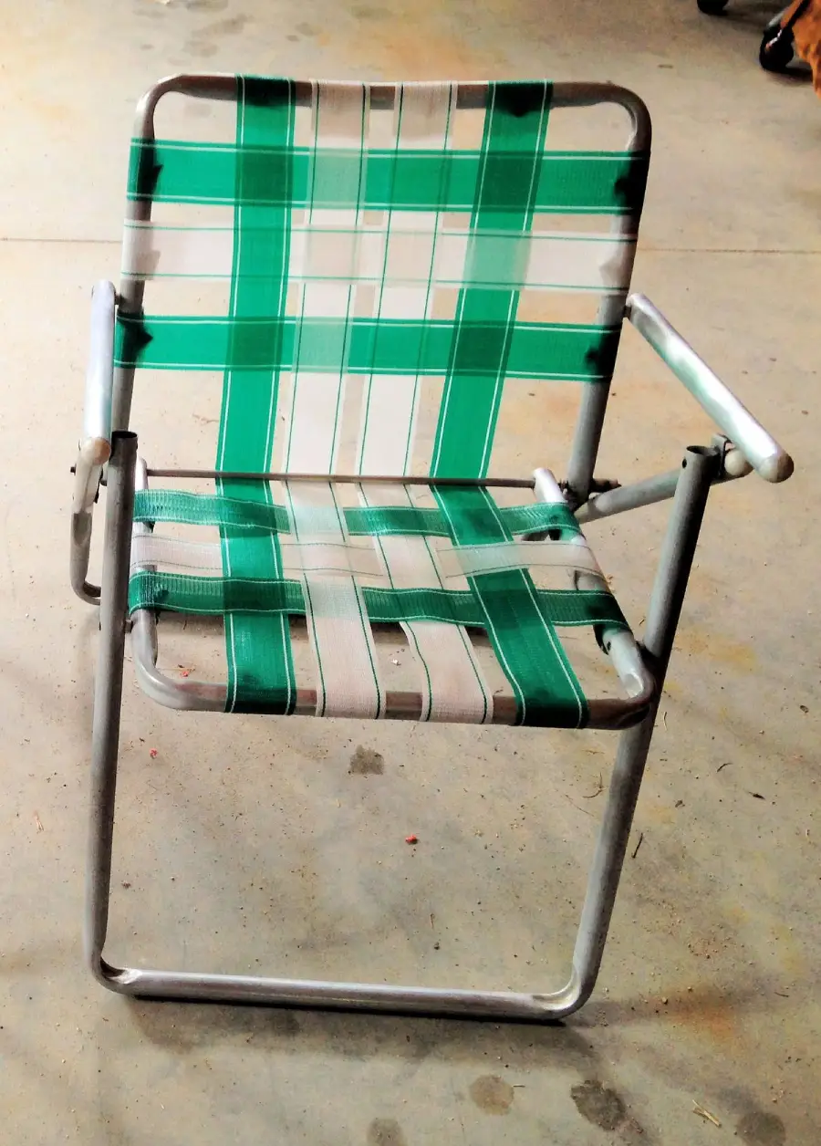 A metal-framed lawn chair with green and white woven plastic strips forming the seat and backrest sits on a concrete floor.