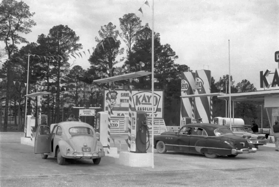 Two vintage cars are parked at a Kay gasoline station with retro pumps, signs, and flags. Pine trees are visible in the background under a cloudy sky.