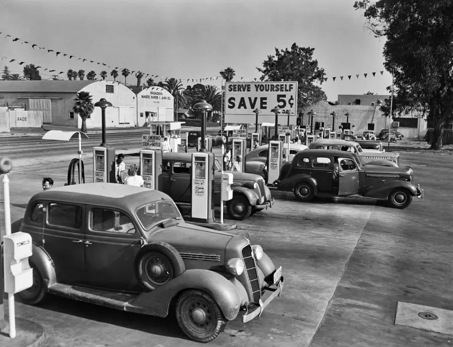 Black and white photo of a vintage gas station with several 1930s cars parked at pumps. A sign reads “Serve Yourself Save 5¢.” People are fueling vehicles; buildings and palm trees are visible in the background.