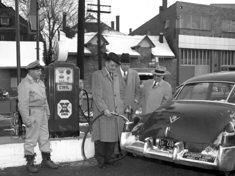 Four men stand at a vintage gas station. One dispenses fuel into a classic car while the others watch. Snow covers some rooftops in the background. The gas pump is labeled “Ethyl.”