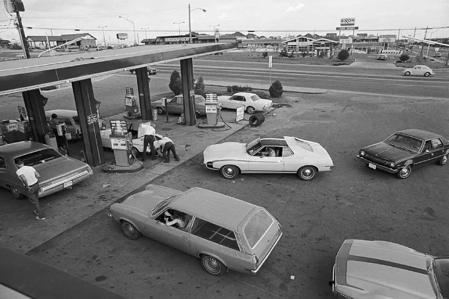 Black-and-white photo of several cars lined up at gas pumps, with people refueling. The scene appears busy, with more cars arriving and businesses visible in the background. The setting suggests a gas shortage era.