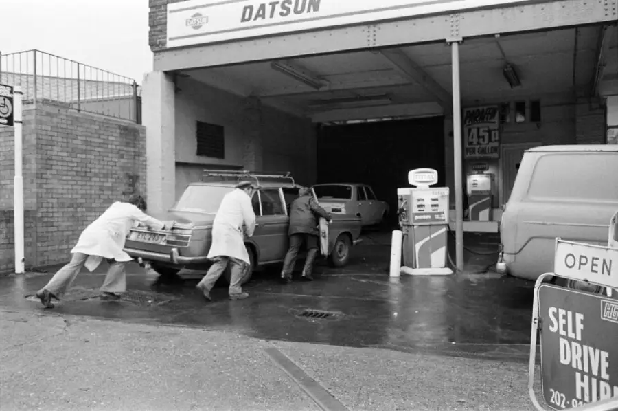 Three people push a car into a vintage gas station garage with a Datsun sign, beside old gas pumps and another parked vehicle. A “Self Drive” sign is visible in the foreground.