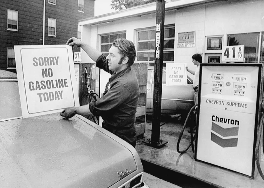 A man at a gas station posts a sign reading “SORRY NO GASOLINE TODAY” on a car during a fuel shortage. Gas pumps and price signs are visible in the background.
