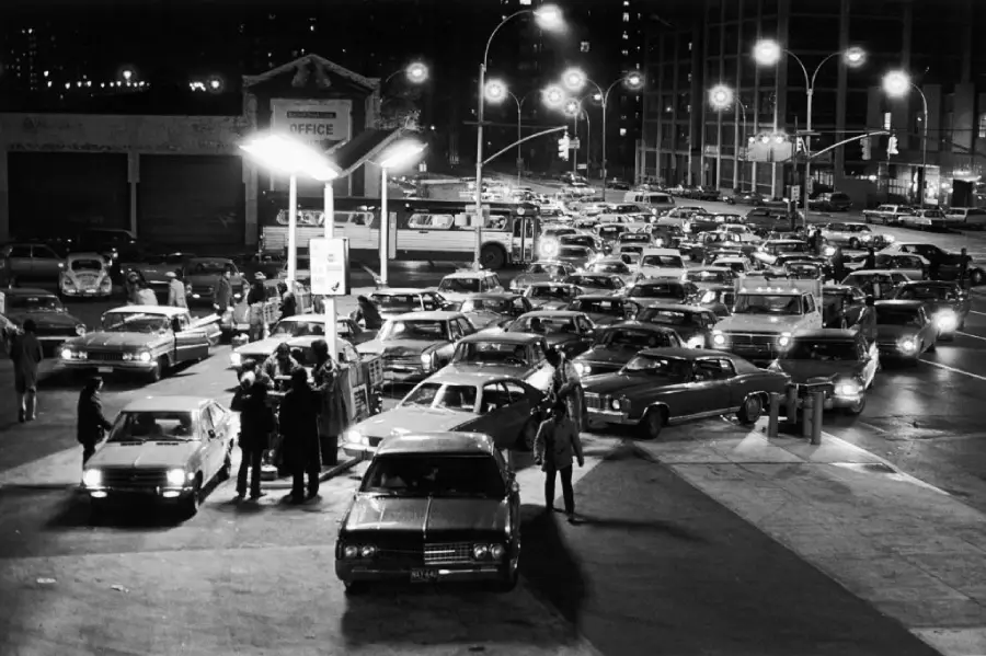 A crowded gas station at night in a city, with lines of cars waiting and people standing near vehicles. Streetlights illuminate the busy urban scene.