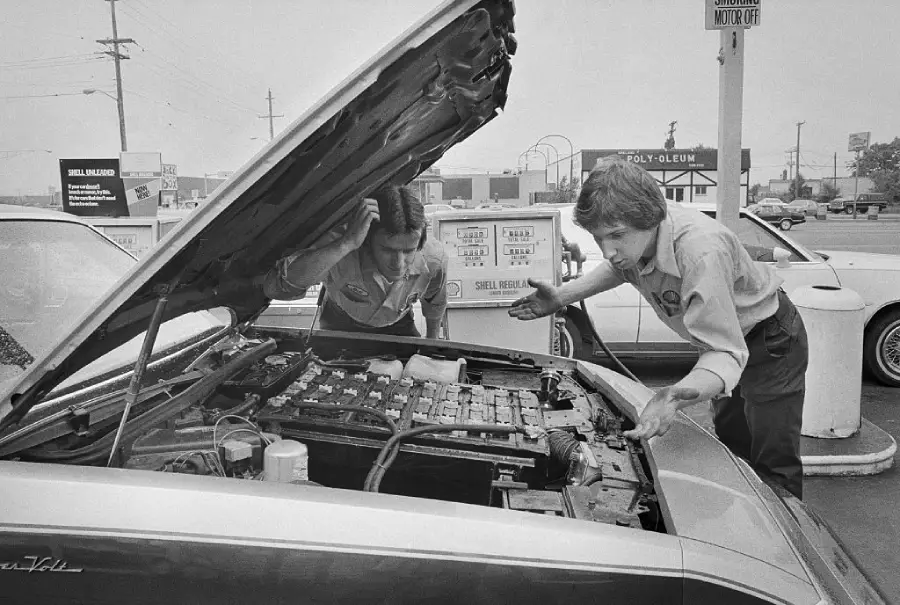 Two mechanics in uniforms look puzzled at the open hood of a car at a gas station, examining the engine area. Signs and cars are visible in the background.