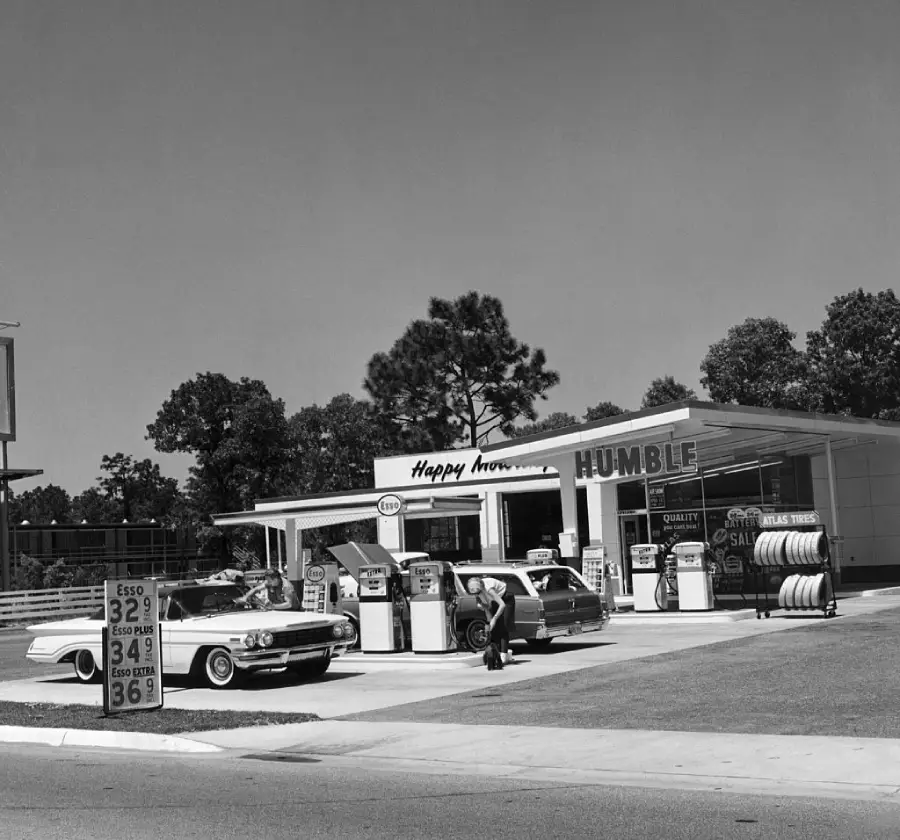 Black and white photo of a vintage gas station with classic cars at pumps, a sign reading “HUMBLE” and gas prices posted. People stand near the cars, and trees are visible in the background.