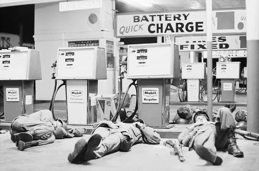 Four soldiers lie on the ground in front of gas pumps at a station, with signs for battery charging and tire fixing visible in the background. Rifles and helmets are on the ground next to them.