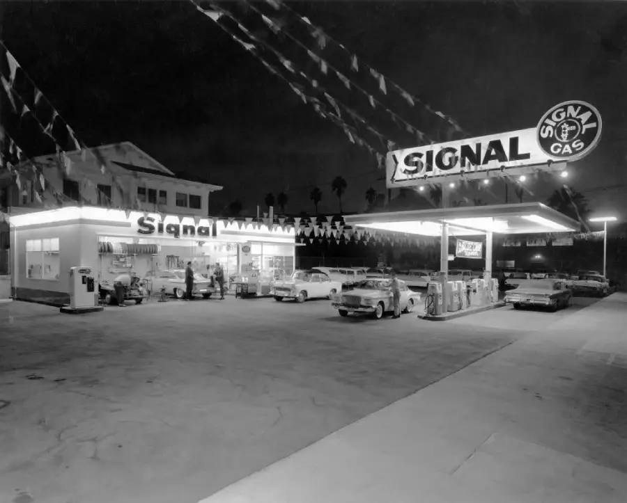 Black-and-white photo of a vintage Signal gas station at night, with lit signs, old-fashioned cars at pumps, and bunting overhead. A building is behind the station, and palm trees are visible in the background.