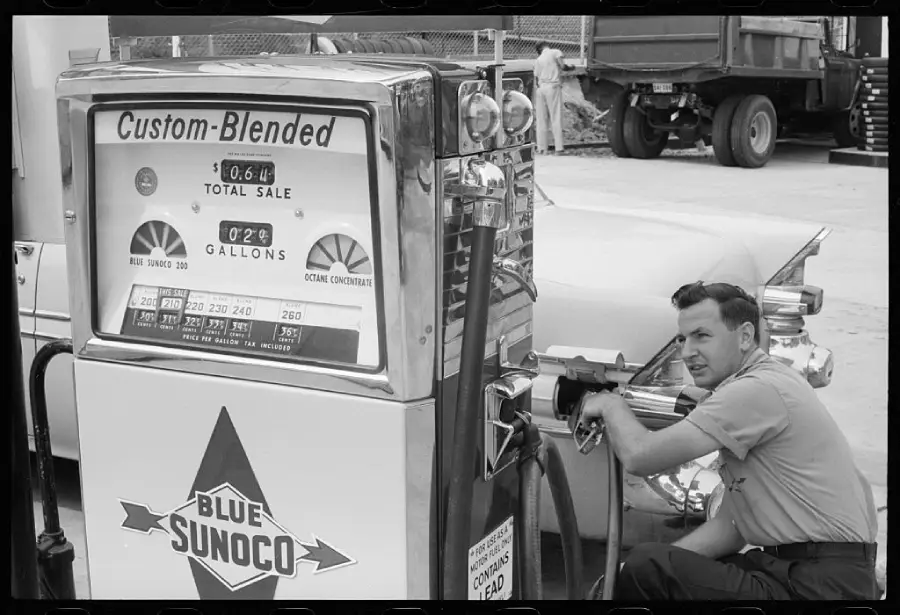 A man kneels beside a vintage Blue Sunoco gas pump, refueling a car at a service station. The pump displays dials, price, and octane levels. A truck and other equipment are visible in the background.