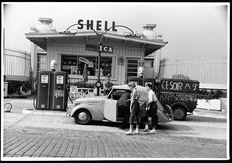 Black-and-white photo of a vintage Shell gas station with old fuel pumps. Three people stand by a classic convertible car. A bicycle and a signboard are visible near the building.