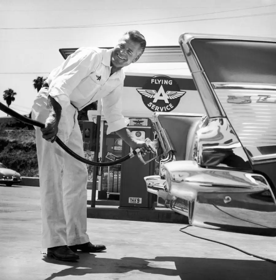 A smiling gas station attendant in uniform pumps fuel into a classic car at a Flying A Service station, with vintage gas pumps and palm trees visible in the background.