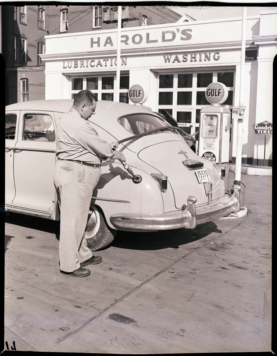 A man fills up a classic car at a vintage Gulf gas station labeled “HAROLD’S LUBRICATION WASHING.” The station features retro gas pumps and a Goodrich sign in the background.