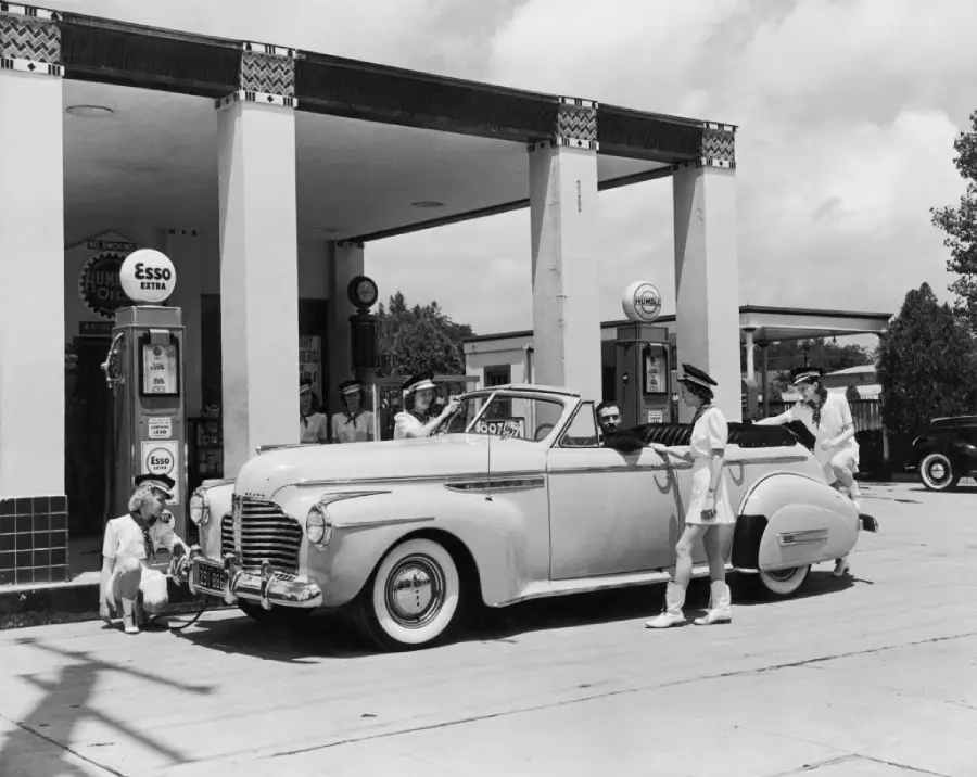 A vintage convertible car with four attendants in white uniforms at an old-fashioned gas station; a child crouches near a front tire while the station building and gas pumps are visible in the background.